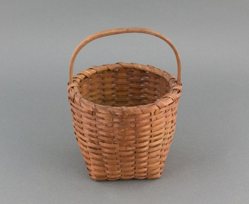Small, ash splint gathering basket with carved handle. Square bottom, vertical sides, and round rim. Ash splint wicker weave with a checker weave base. The inner rim is carved wood with an outer, splint rim held in place with a single ash splint lash.